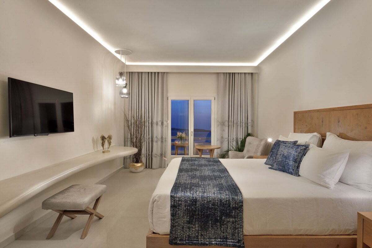 Una habitación de hotel moderna con una cama grande, un televisor, una mesita con una silla y vistas al mar a través de las ventanas.