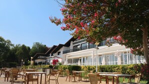 An outdoor seating area with wicker tables and chairs, a white building with large windows, and a tree with pink flowers. - Hotel Gudamendi (San Sebastián)