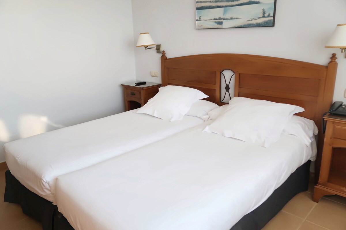 A double bed with white linens, a wooden headboard, two bedside tables, and a wall-mounted lamp.
