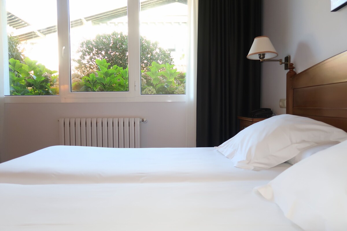 A hotel room with a bed, a window with curtains, a radiator, and a bedside lamp.