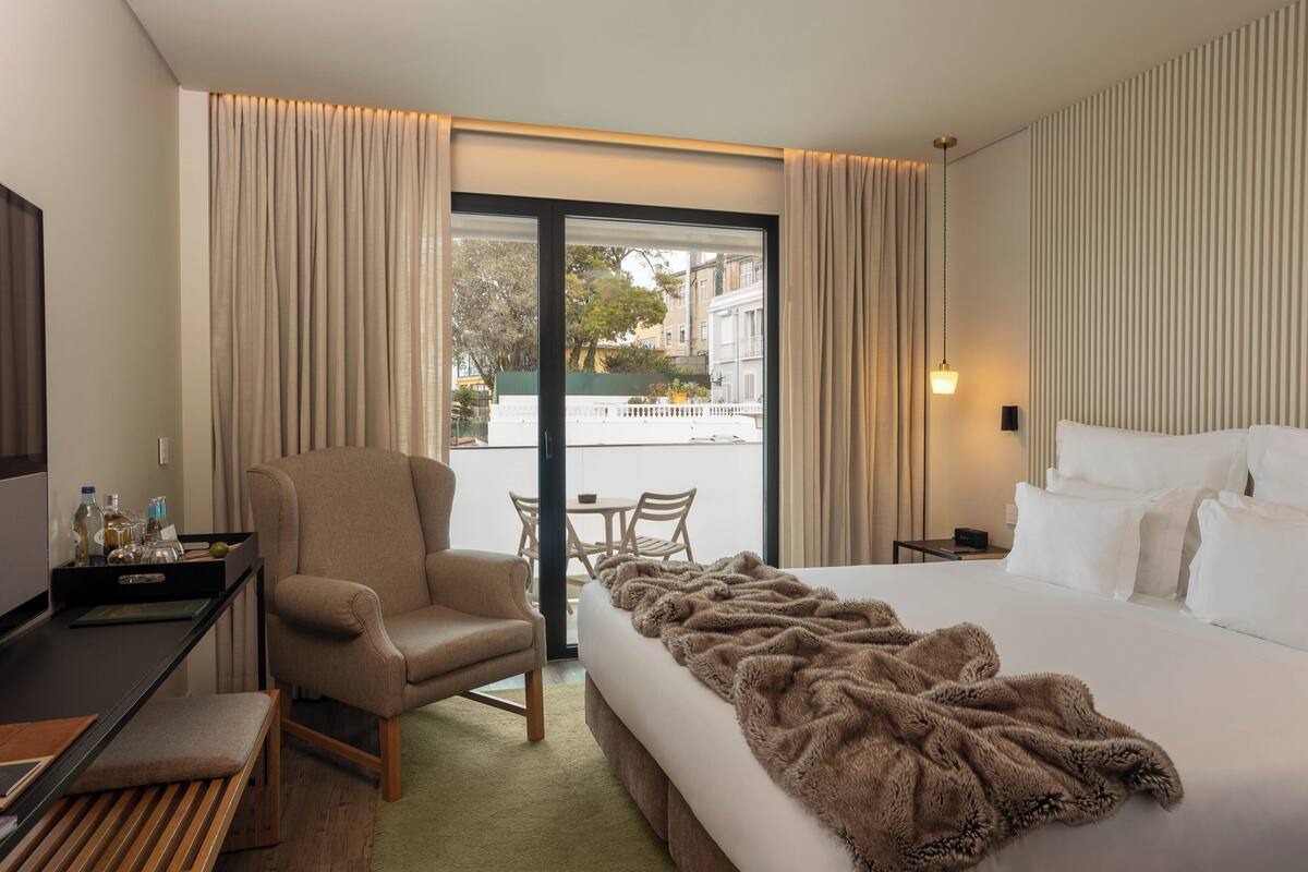 A hotel room with a large bed, a chair, a desk, and a balcony with a view of buildings and trees.