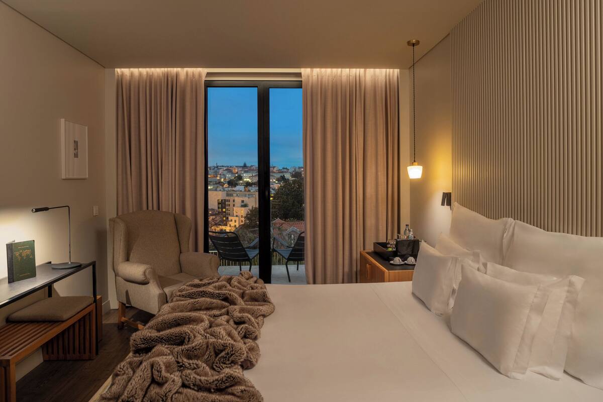 A hotel room with a large bed, a chair, a desk with a lamp, and a view of the city through the window.