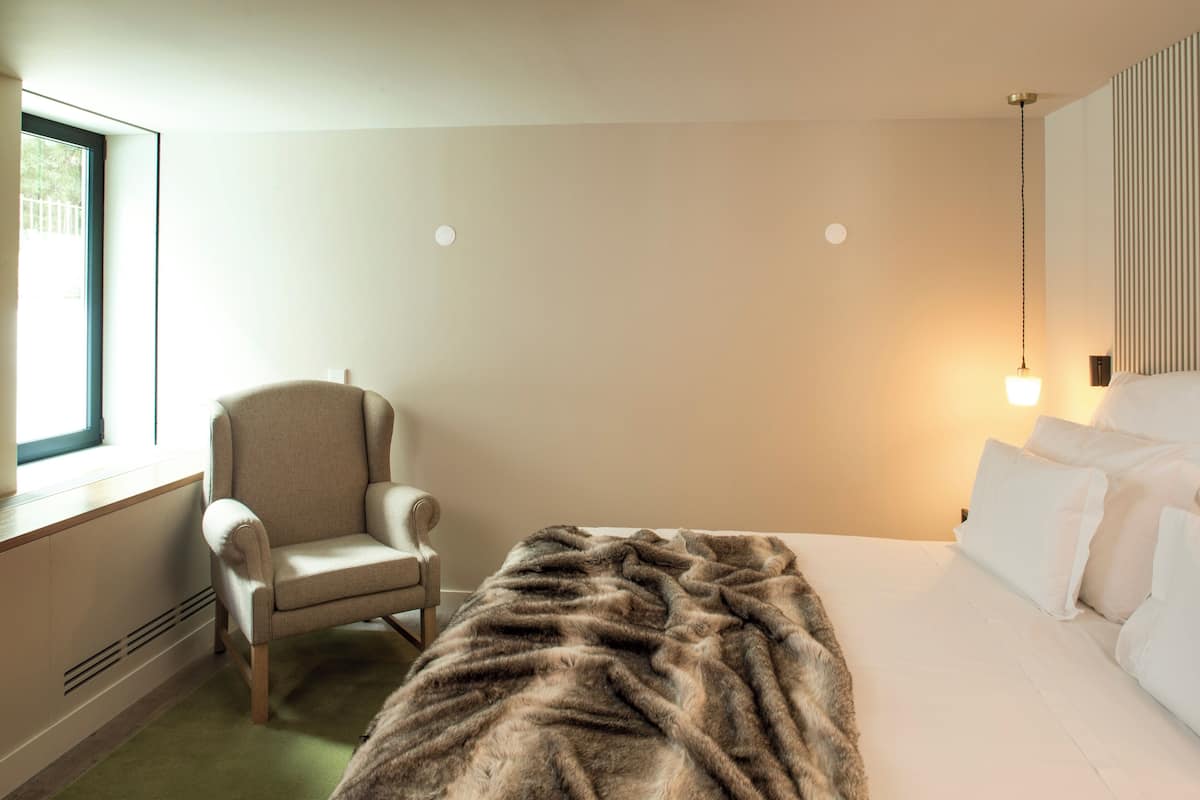 A hotel room with a bed, a chair, a window, and a wall-mounted light fixture.