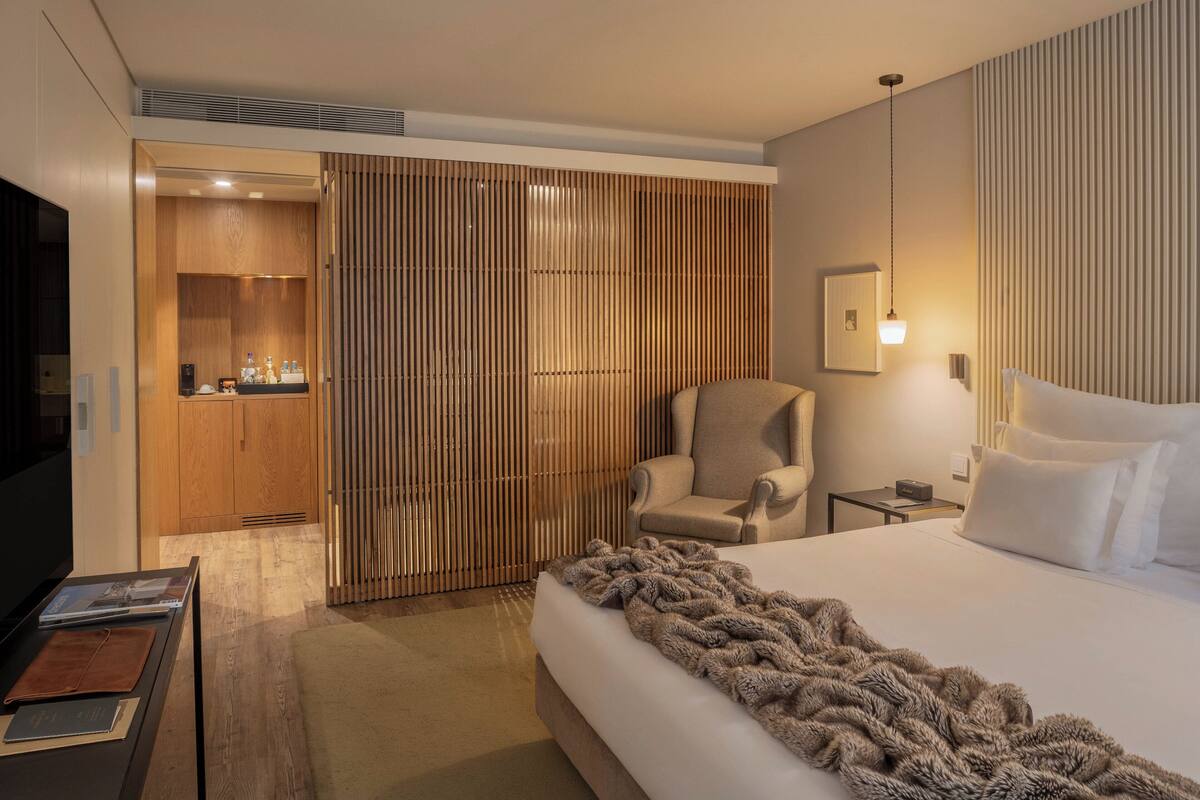 A modern hotel room with a large bed, a seating area with a chair, a TV, and a wooden wardrobe.