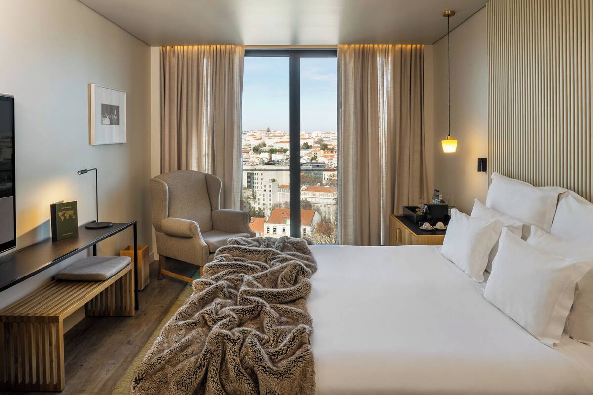 A hotel room with a large bed, a desk, a chair, and a view of a cityscape through a large window.