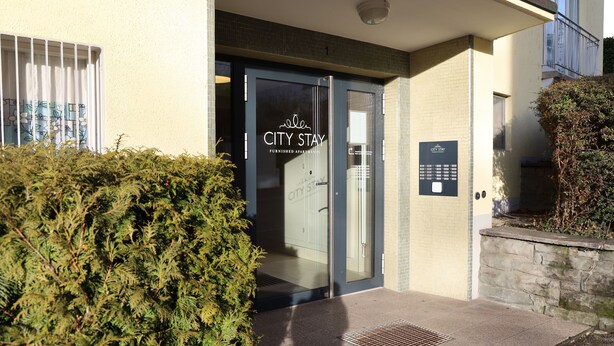 A building entrance with a glass door, a sign indicating 'City Stay Furnished Apartments', and a small stone wall with a plant. - City Stay Apartments Forchstraße (Zürich)