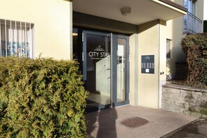 A building entrance with a glass door, a sign indicating 'City Stay Furnished Apartments', and a small stone wall with a plant. - City Stay - Forchstrasse (Zürich)