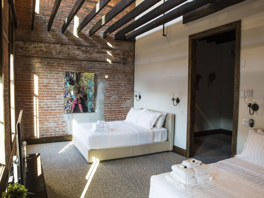 Quarto de hotel com parede de tijolos, uma cama grande, uma cama menor e um quadro na parede.