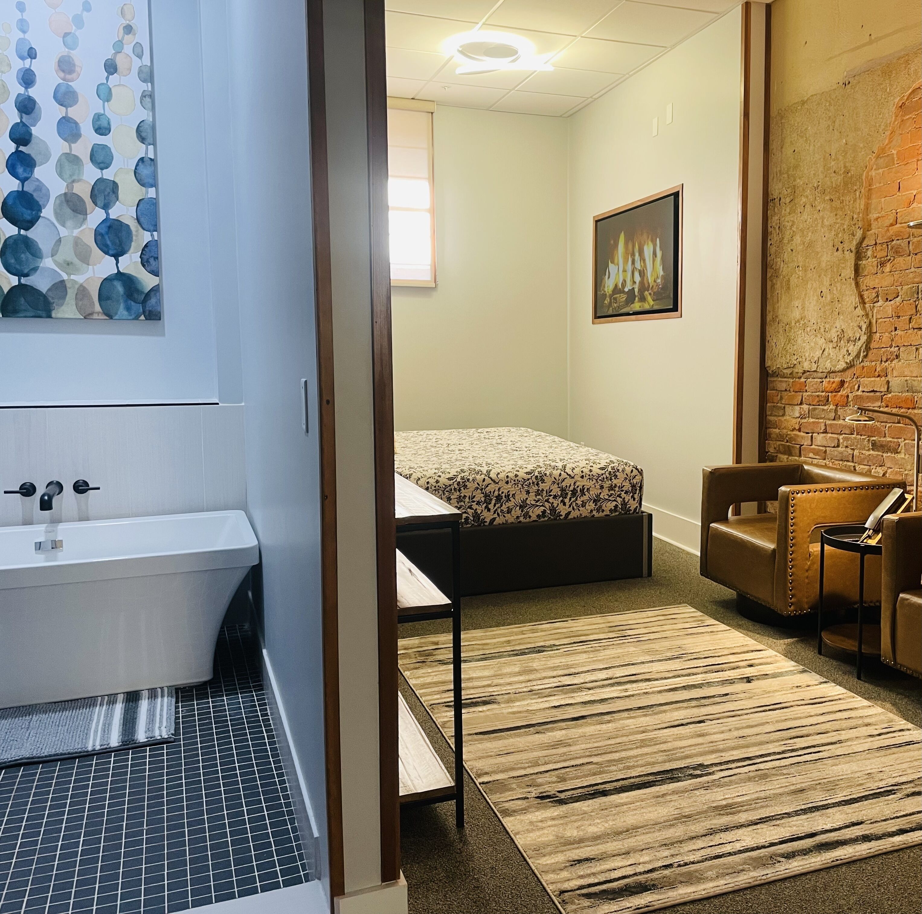Une chambre d’hôtel moderne comprenant un lit, une petite table, une chaise, une salle de bain avec baignoire et un mur en briques.