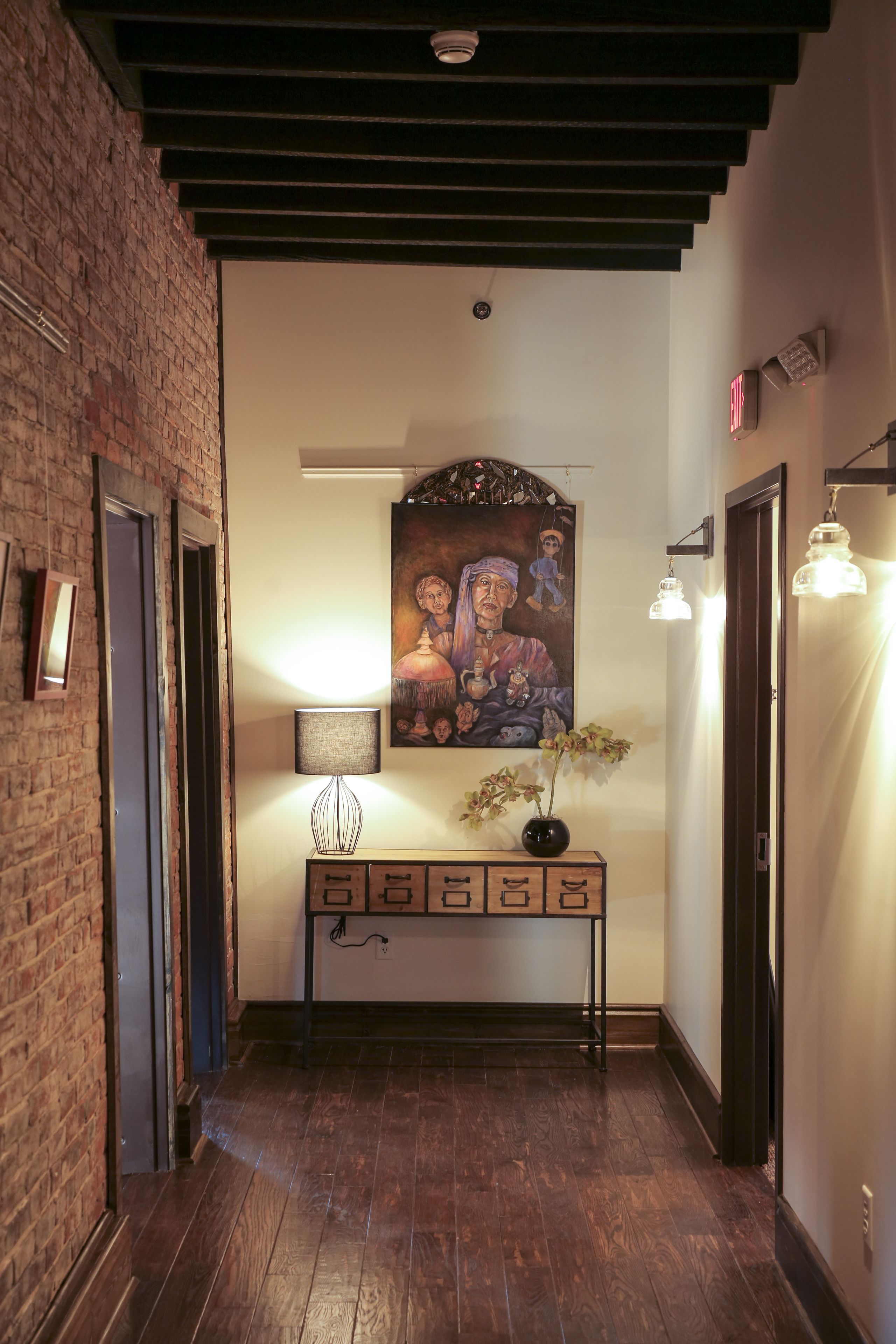 A hallway with wooden flooring, a brick wall, a painting, a lamp, and a console table.
