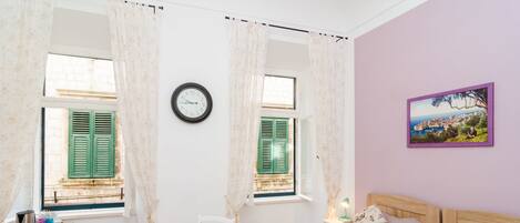A room with two beds, a wooden table, a chair, a clock, and a window with shutters.