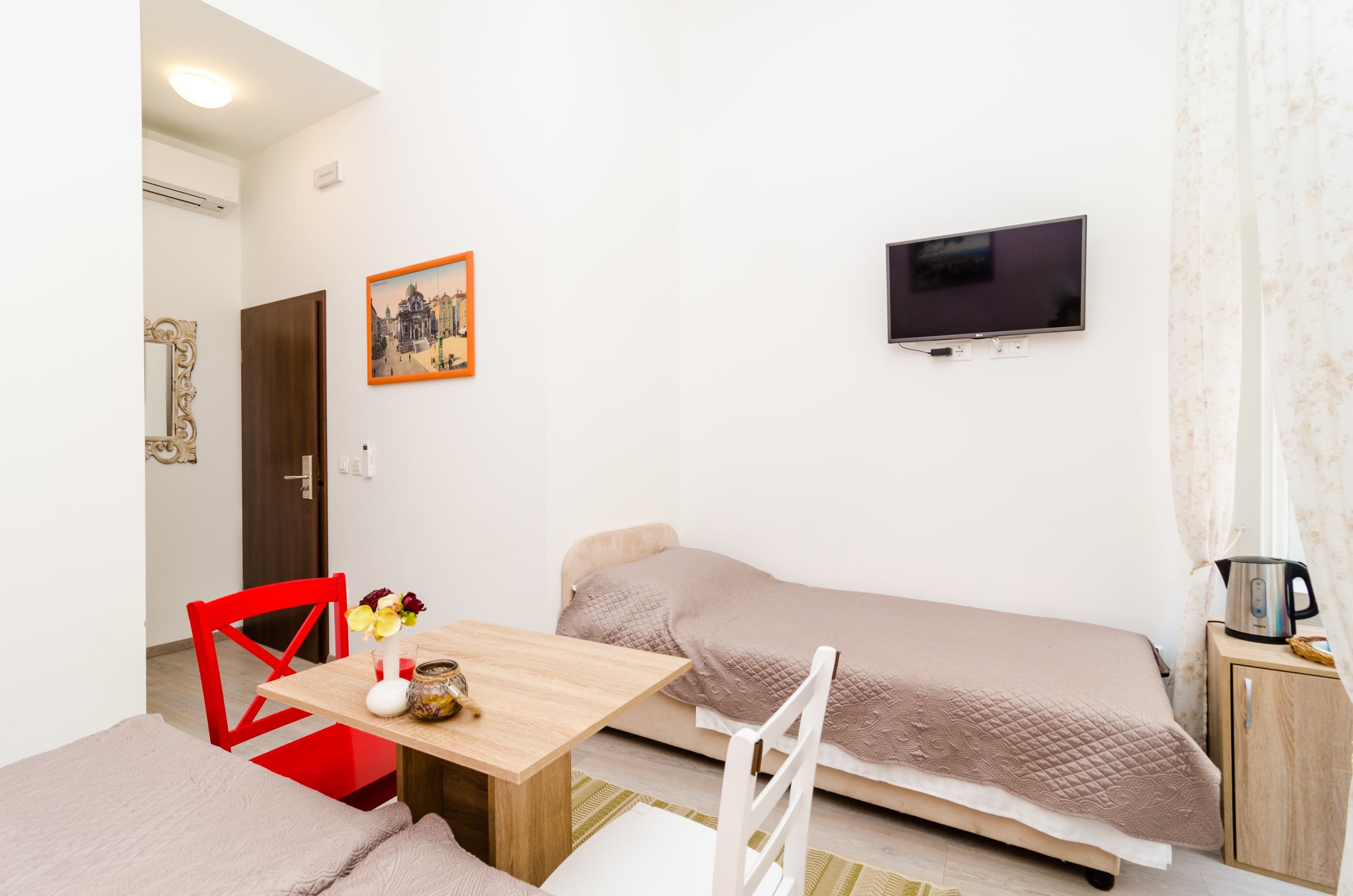 Une pièce de taille modeste comprenant un lit, un téléviseur fixé au mur, une petite table avec un vase et un bol, et une chaise rouge.