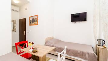 Une pièce de taille modeste comprenant un lit, un téléviseur fixé au mur, une petite table avec un vase et un bol, et une chaise rouge.
