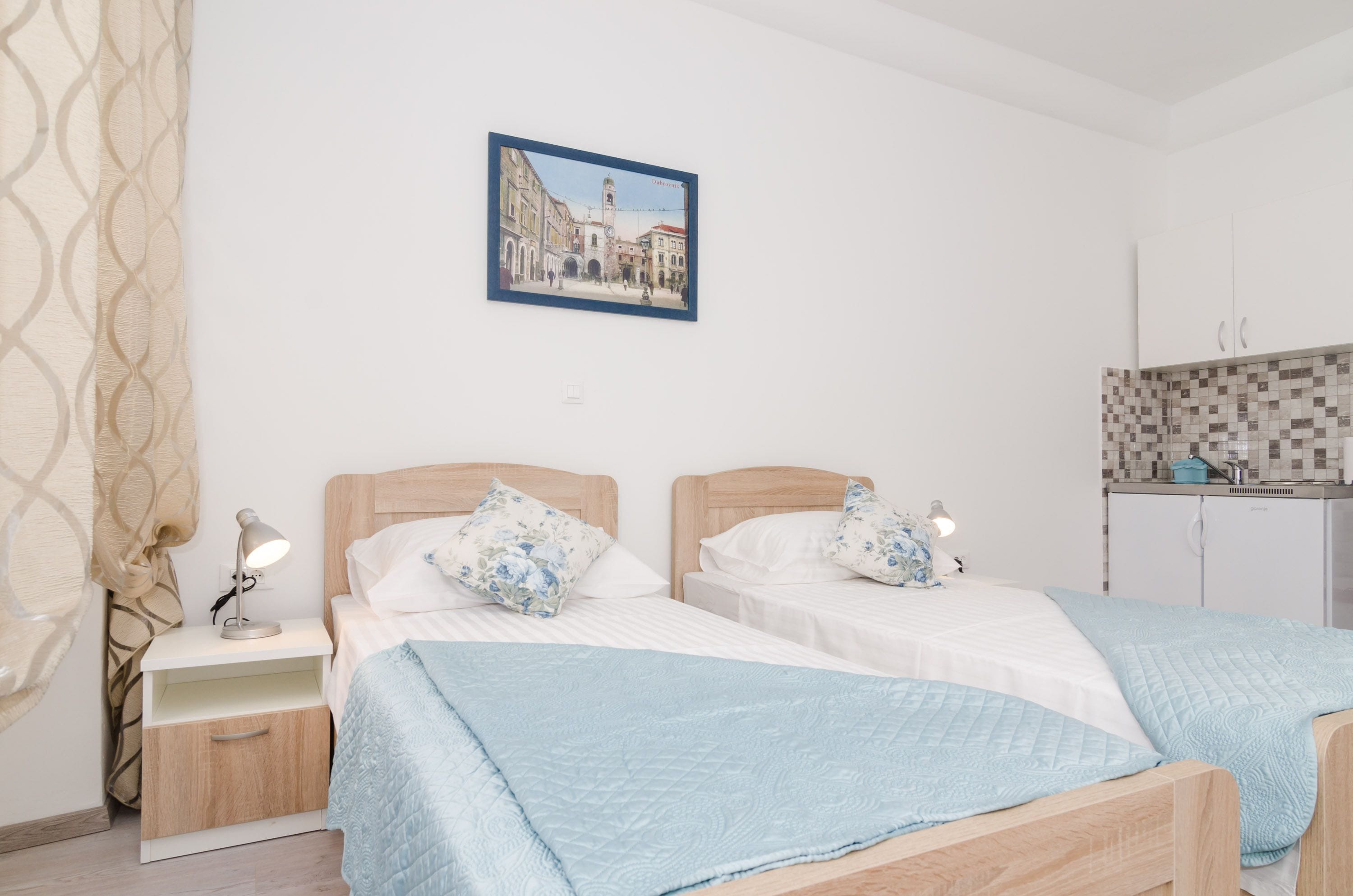 A bedroom with two beds, a nightstand with a lamp, a wall-mounted picture, and a kitchenette area.