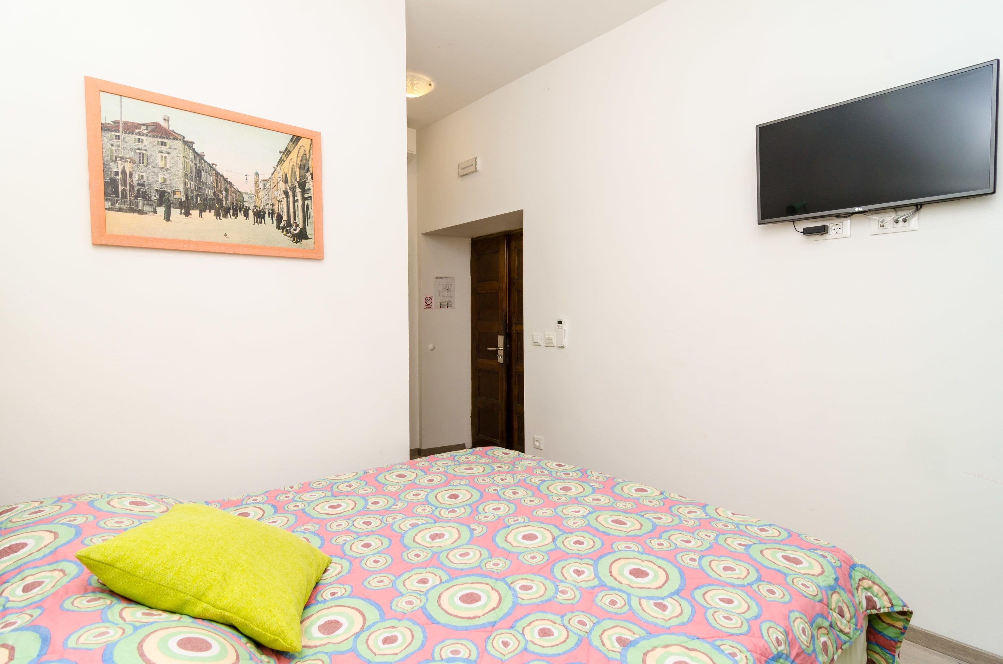 Un dormitorio con una cama, televisión, un cuadro enmarcado y una puerta que da a otra habitación.