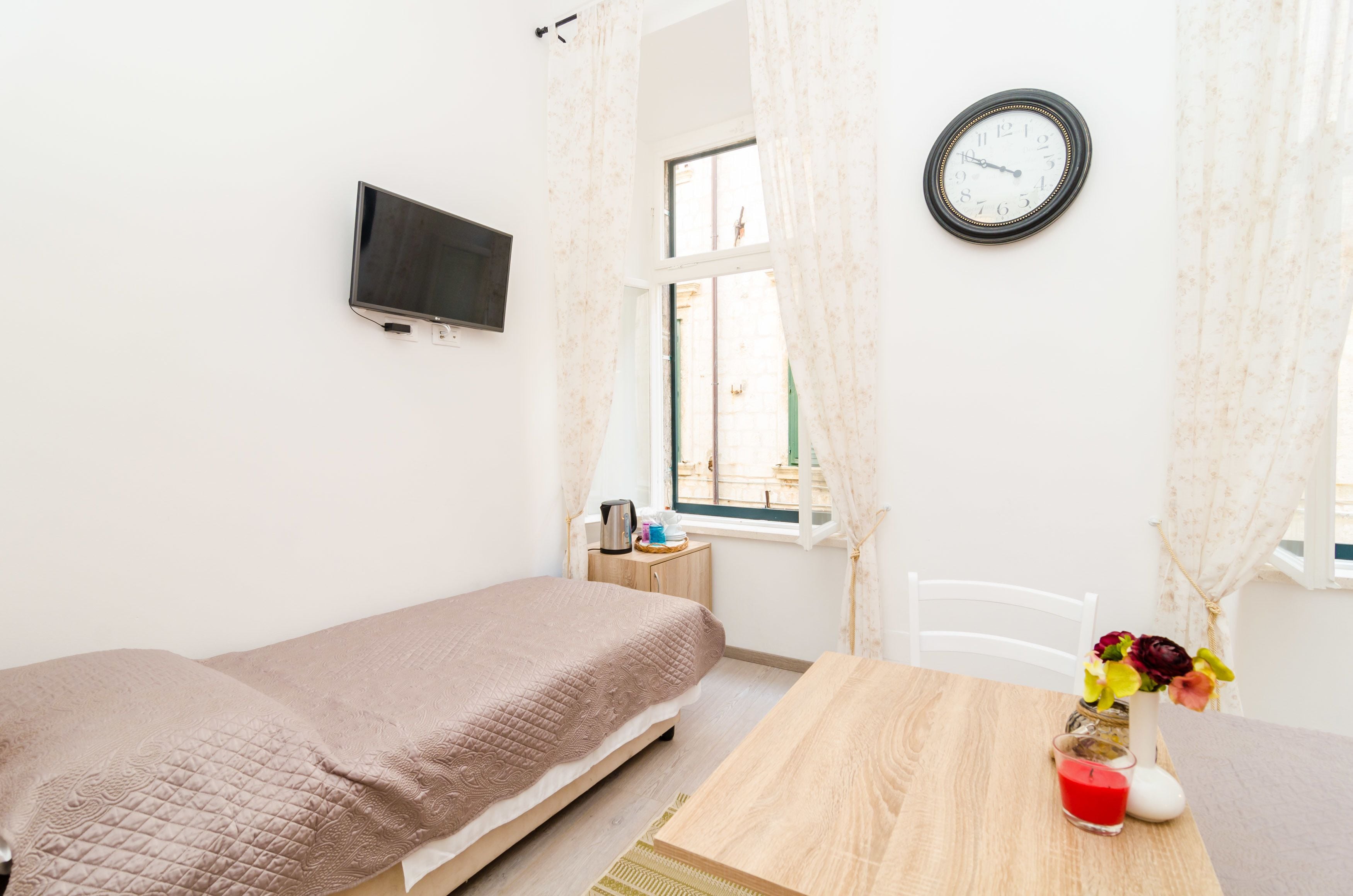 A small, well-lit bedroom with a single bed, a wooden table, a clock, and a TV mounted on the wall.