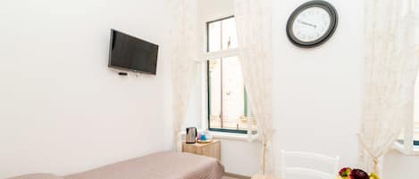 A small, well-lit bedroom with a single bed, a wooden table, a clock, and a TV mounted on the wall.