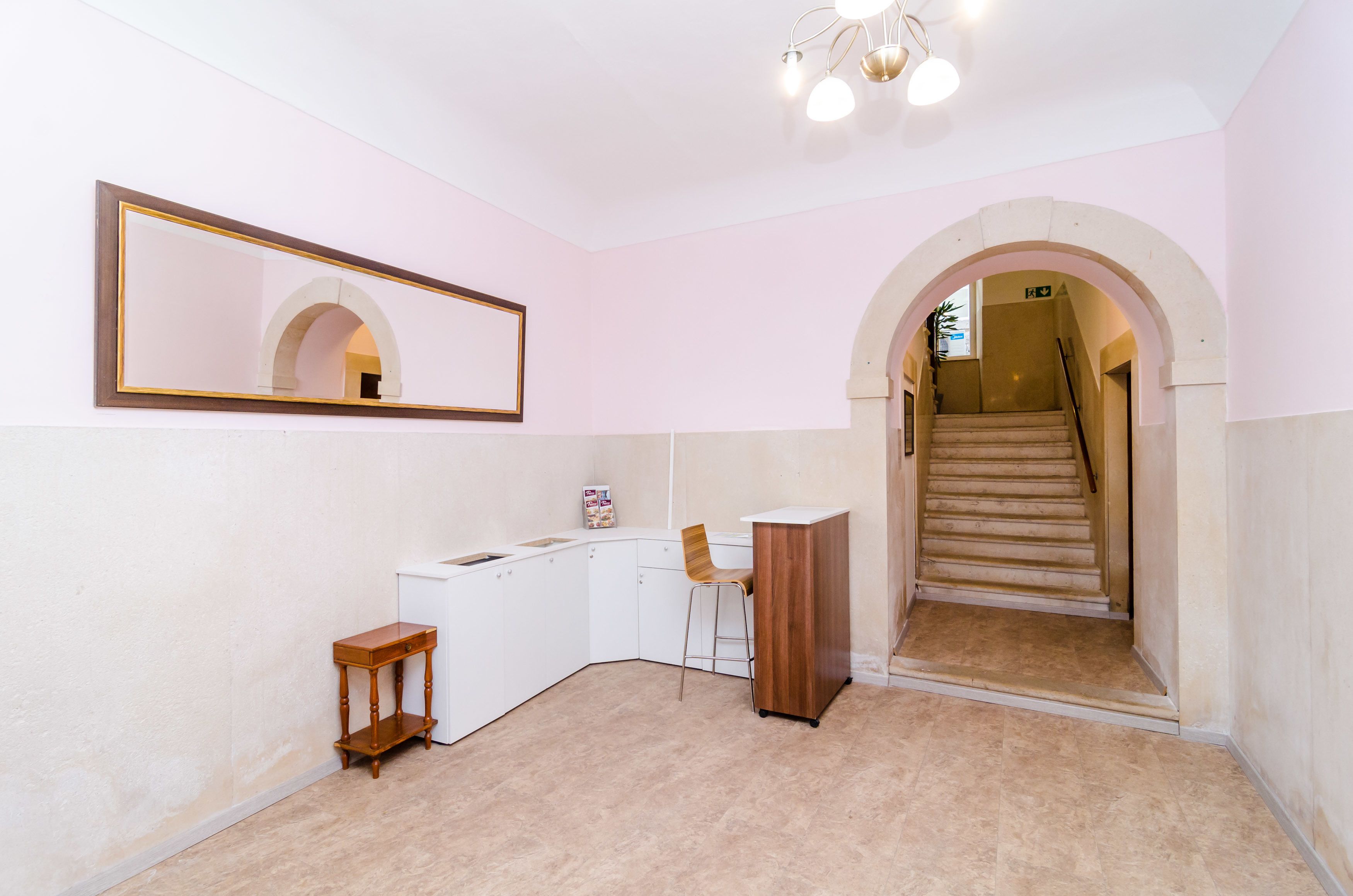 Ein Zimmer mit weißem Schreibtisch, einem Stuhl, einem kleinen Holzschemel, einem Spiegel und einer Treppe, die zu einer anderen Ebene führt.