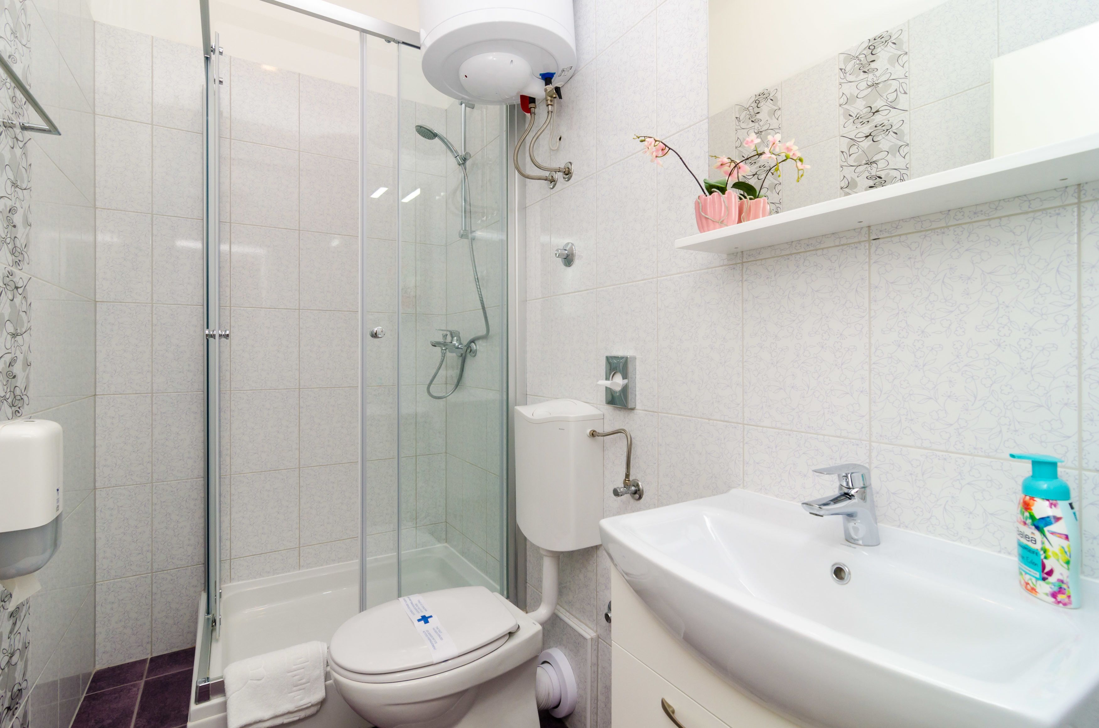Un baño moderno con ducha envuelta en vidrio, inodoro blanco, lavamanos con grifo y dispensador de papel higiénico.
