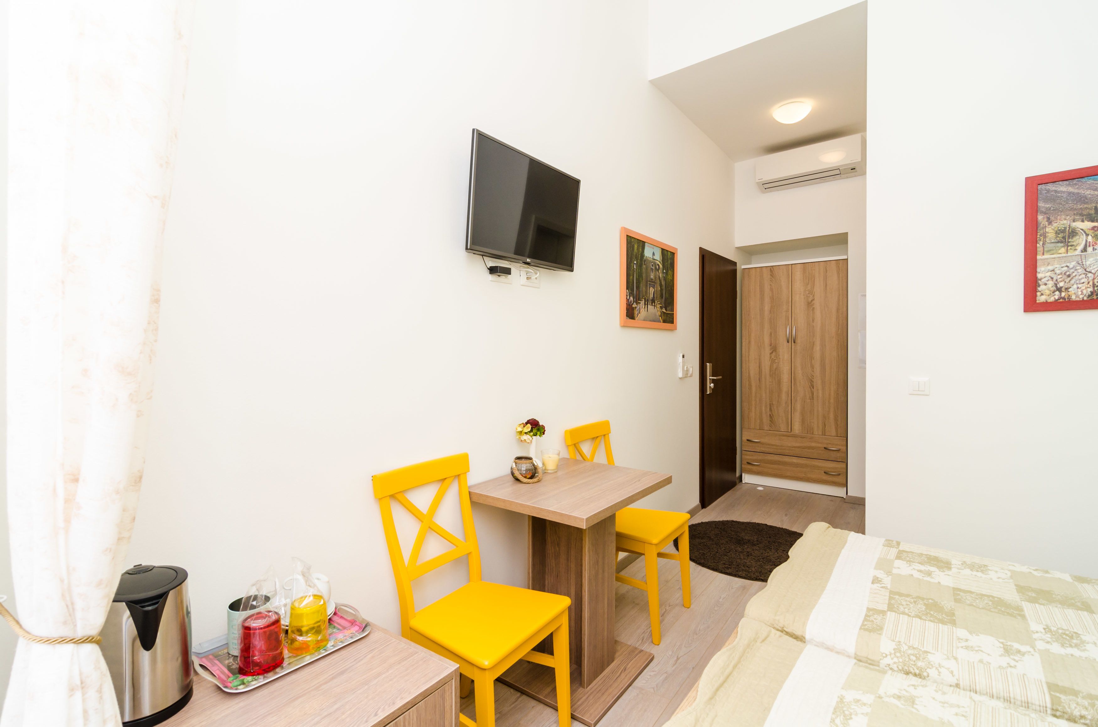 A small, bright room with a wooden table, two yellow chairs, a TV mounted on the wall, and a wooden wardrobe.