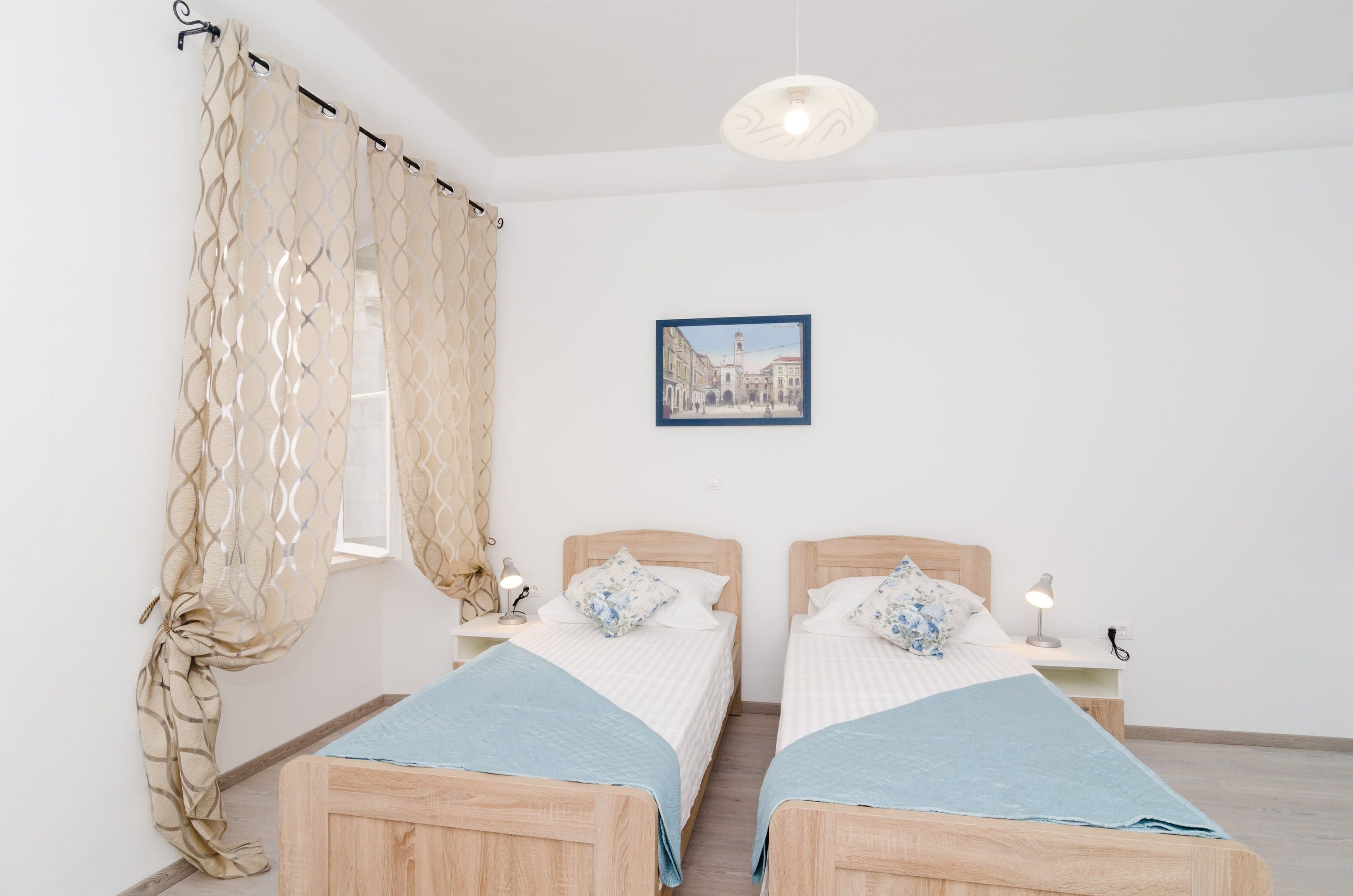 Une chambre à coucher avec deux lits en bois, une fenêtre avec des rideaux, une table de chevet avec une lampe et un tableau encadré au mur.