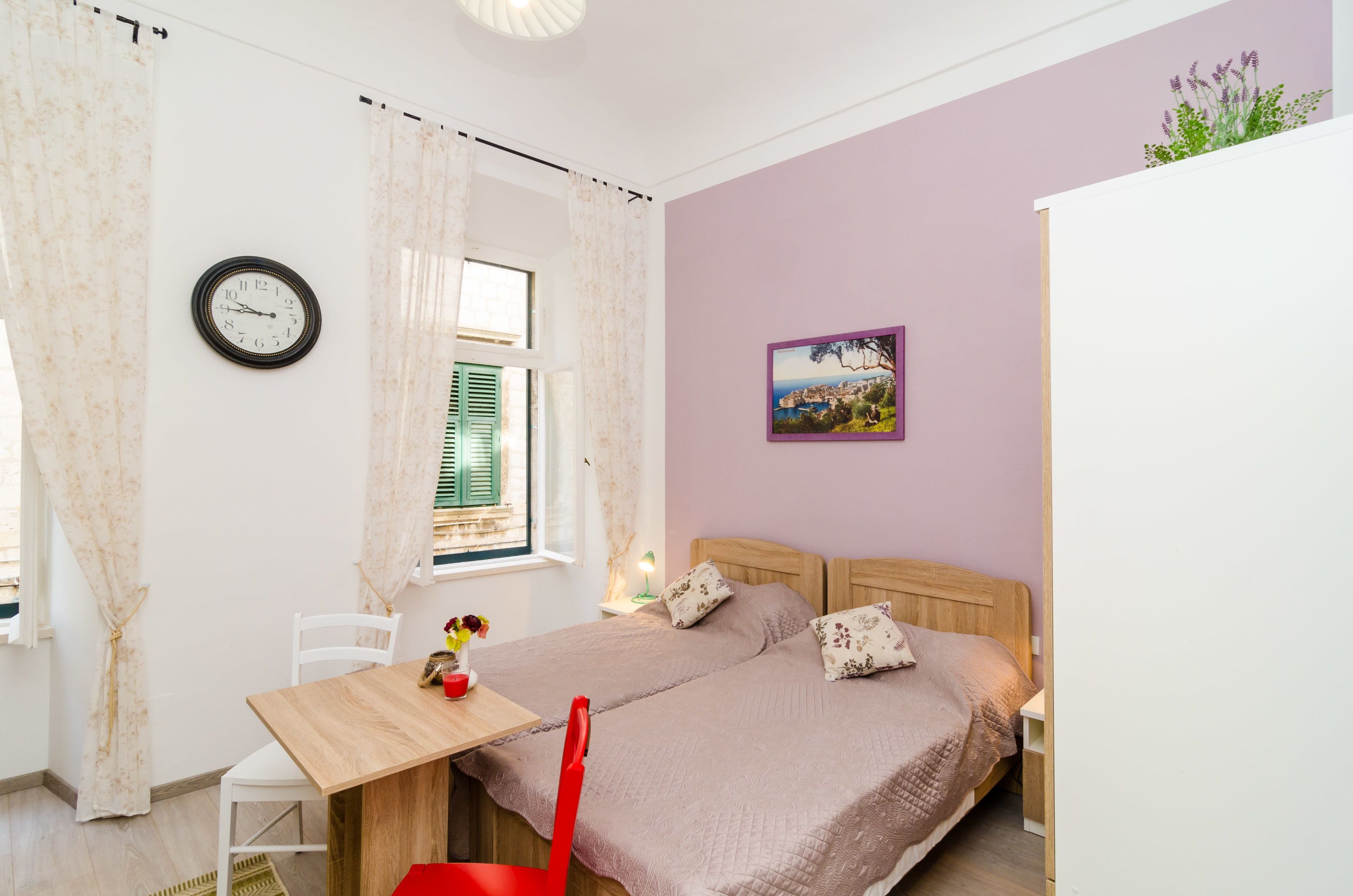 A bedroom with a bed, a wooden table, a chair, a window with curtains, a wall clock, and a picture.