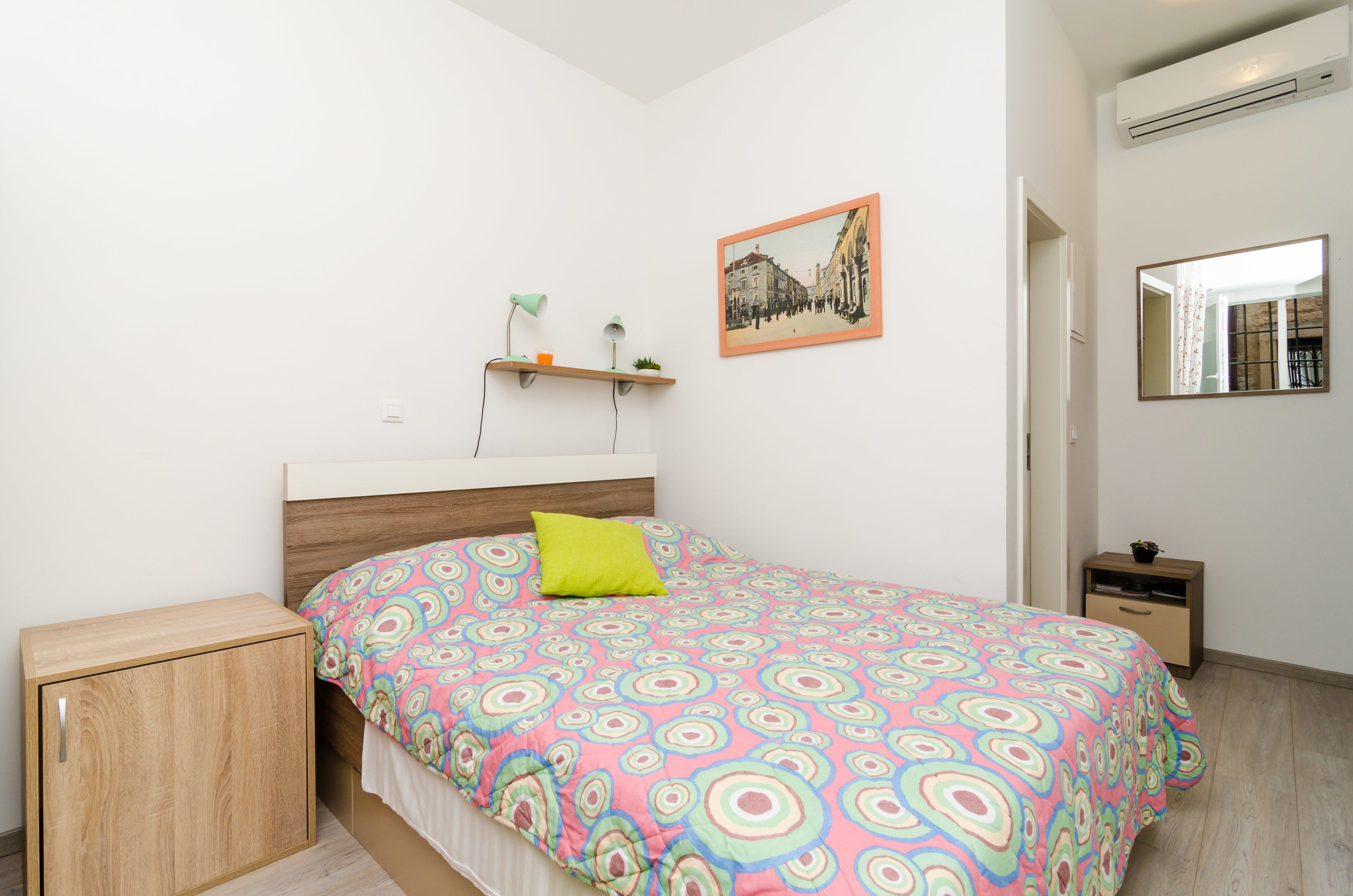Un dormitorio con cama, mesita de noche, una pequeña estantería con una planta, un cuadro en la pared y un espejo.