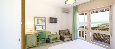 A hotel room with a bed, a desk with a chair, a small table, a trash bin, a framed picture, and a view of the sea through a balcony.