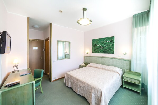A hotel room with a bed, a desk with a lamp, a chair, a nightstand, and a painting on the wall.