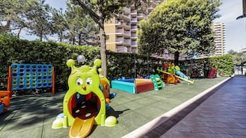Children can play on a slide, climbing frames, and a large dinosaur figure in this outdoor play area surrounded by hedges and trees.