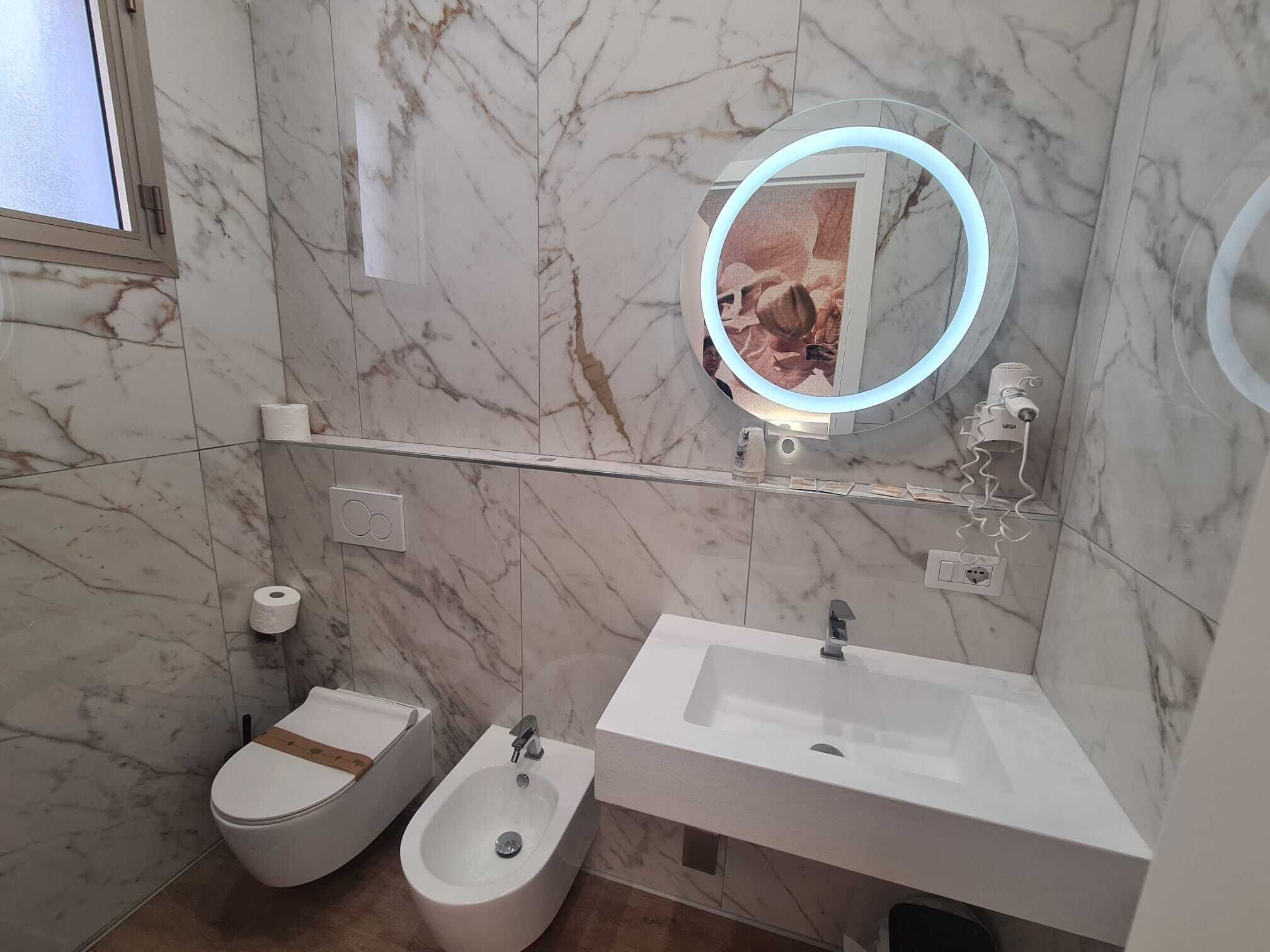 A bathroom with a bidet, a sink, a mirror with a light, and marble walls.