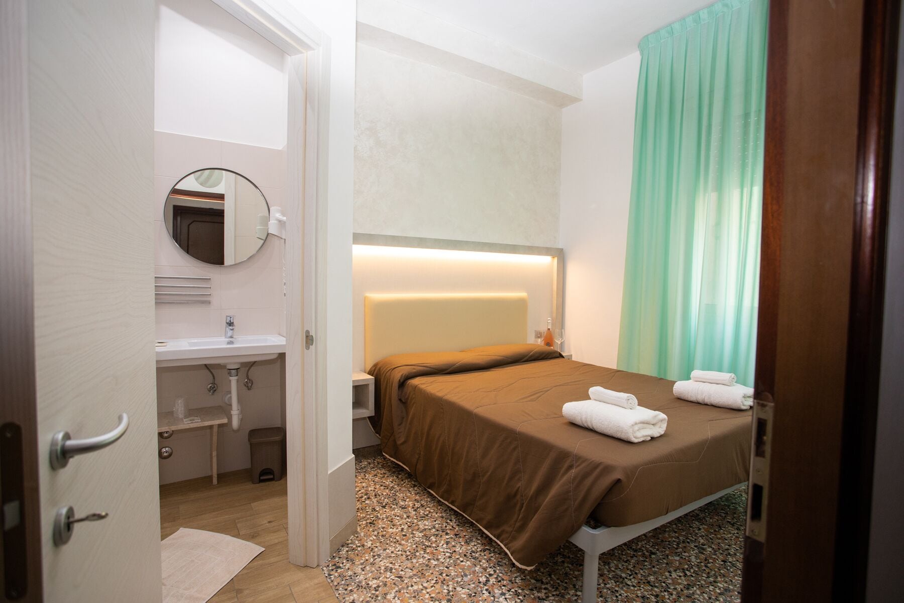 Quarto de hotel com cama, mesa de cabeceira, espelho, pia e janela com cortinas.