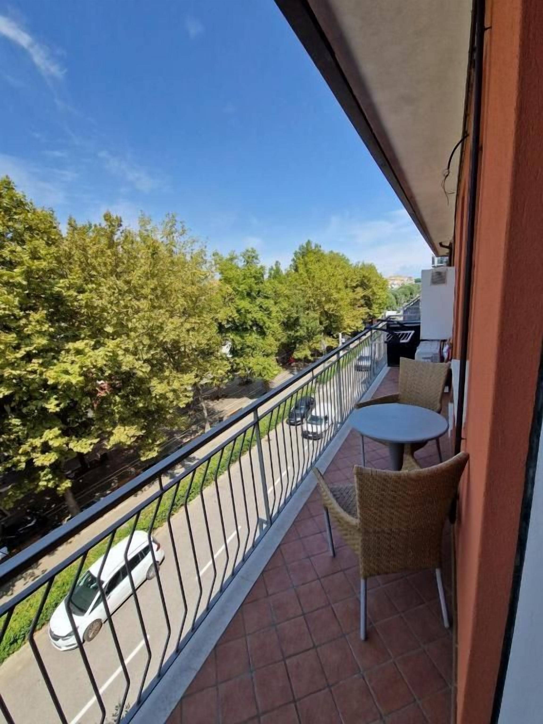 Varanda com vista para a estrada e árvores, uma mesa pequena e uma cadeira.