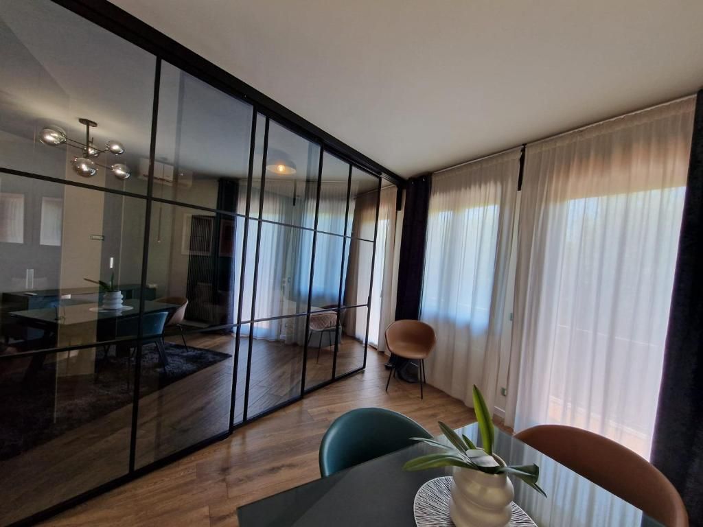 A modern living room with a glass partition, a dining area with a table and chairs, and a large window with curtains.