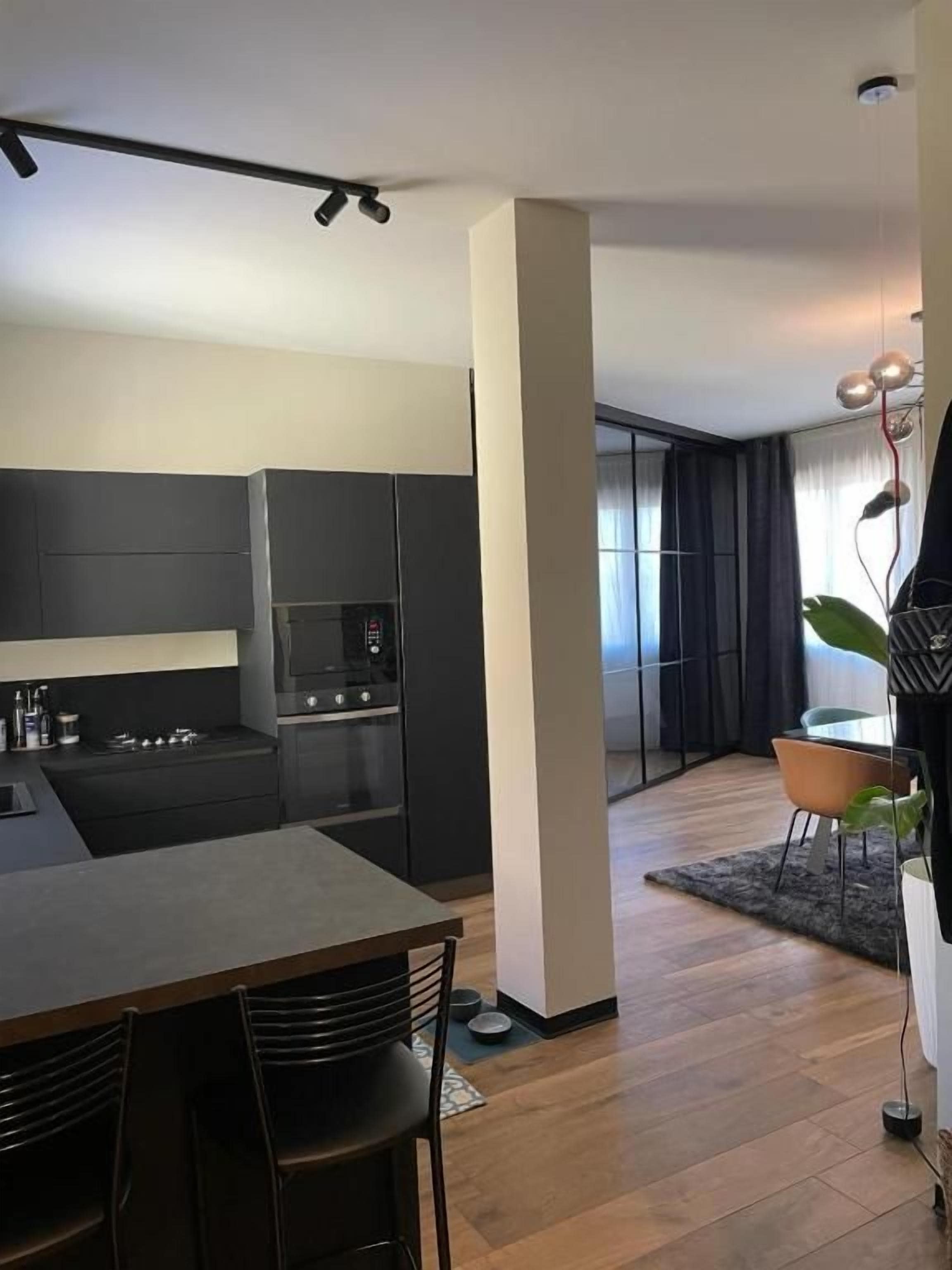 Una cocina moderna con armarios oscuros, un comedor con mesa y sillas, y vistas hacia una sala de estar con un amplio ventanal.