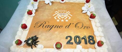 Un gâteau rectangulaire à la couleur dorée, décoré de fleurs blanches et de fraises, sur lequel on peut lire l’inscription « HÔTEL Ragno d’Oro 2018 ».