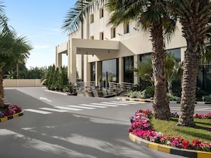Un edificio moderno con ingresso coperto, palme e aiuole fiorite.