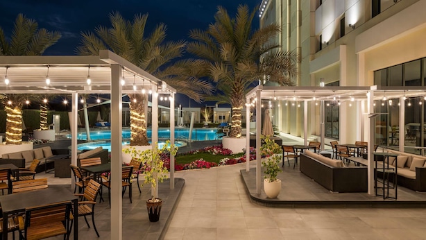 An outdoor pool area with seating and string lights, surrounded by palm trees and a modern building.