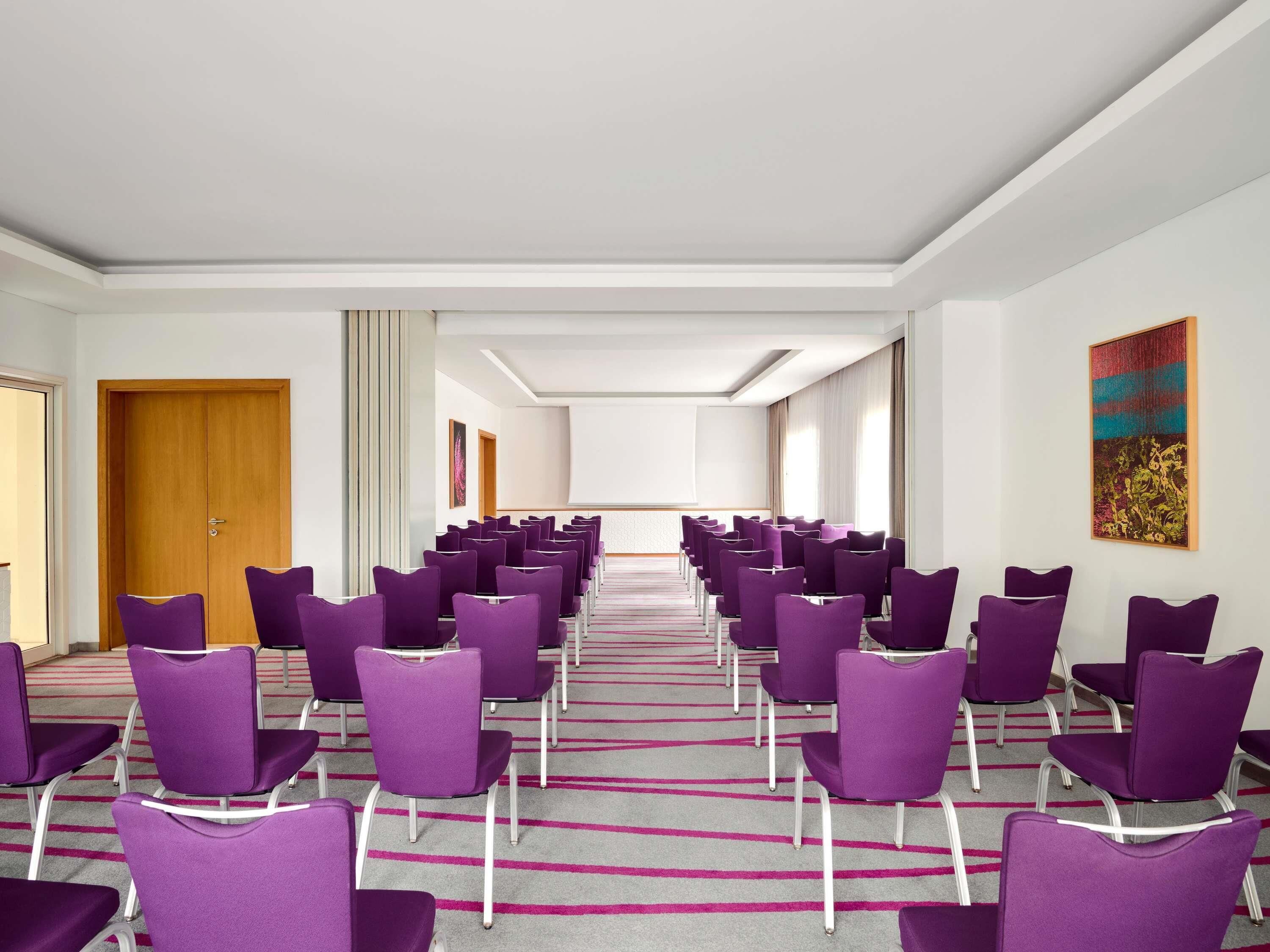 A conference room with rows of purple chairs, a carpeted floor, a wooden door, and a painting on the wall.
