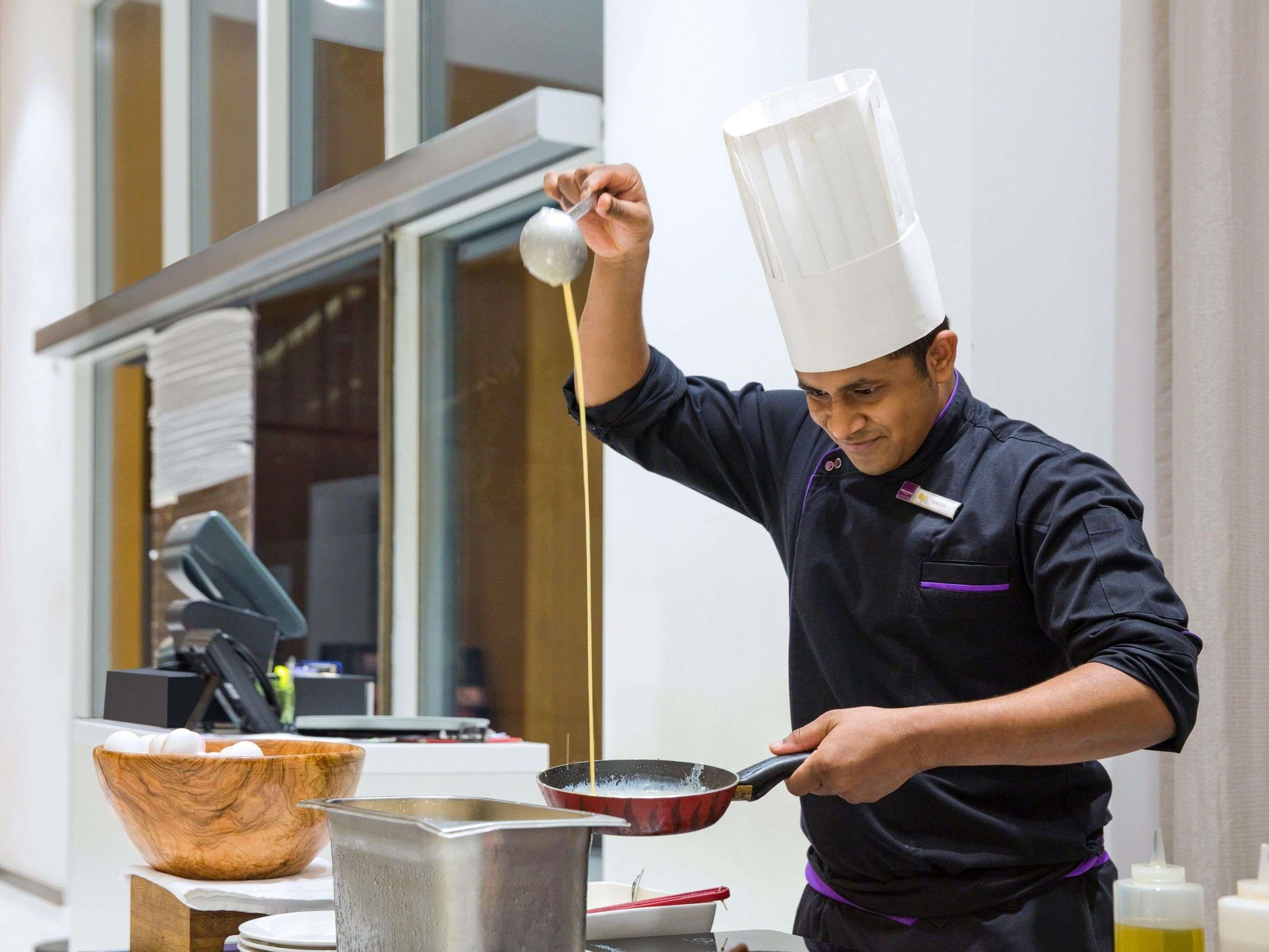 A chef in a professional kitchen, holding a ladle over a pan, with a bowl of eggs and a pot on the counter.