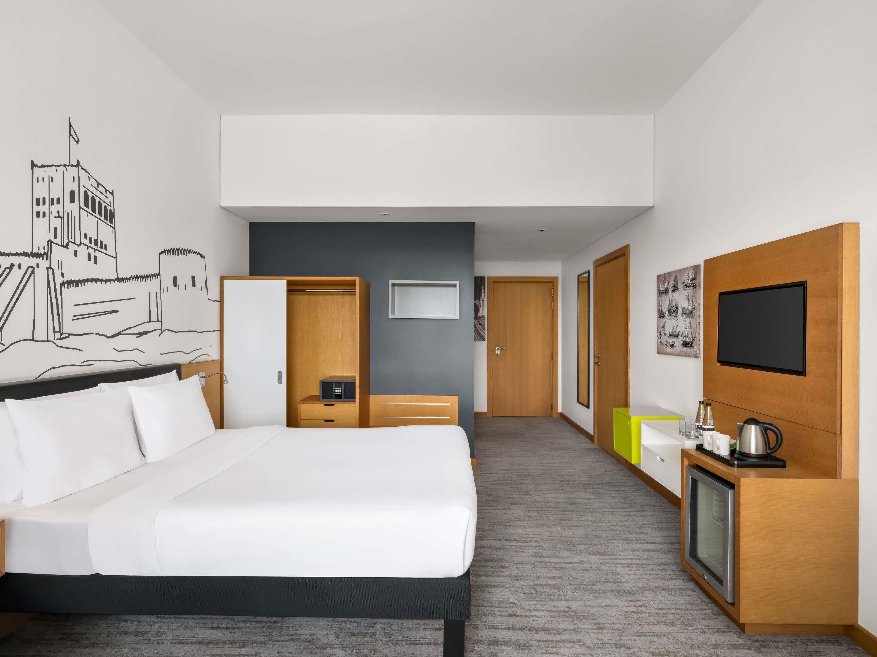 Quarto de hotel com cama, guarda-roupa, secretária com chaleira, televisão e parede com um mural de paisagem urbana.