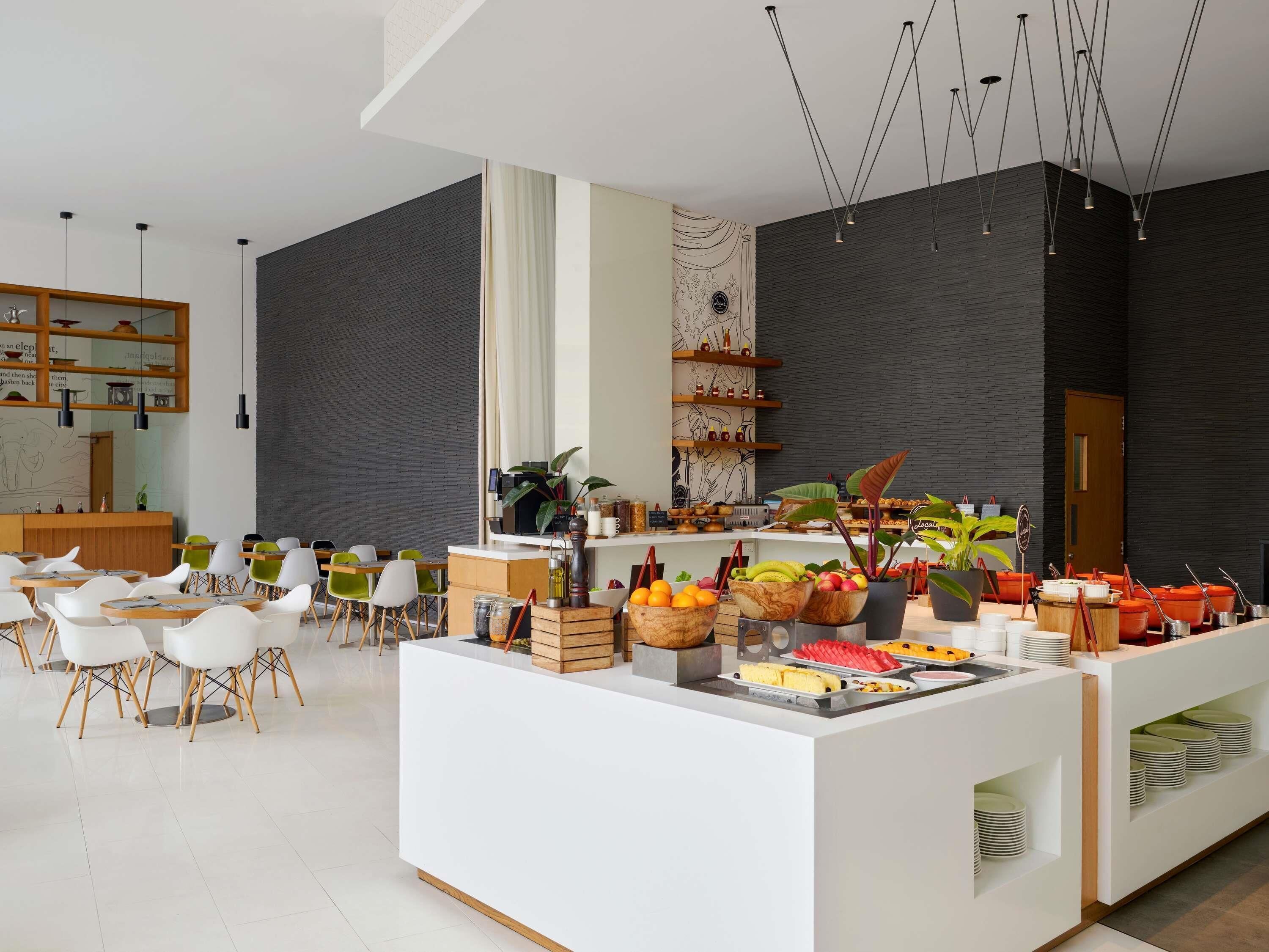 Un espace d’accueil moderne avec un comptoir de réception blanc, des chaises en bois et un buffet proposant des fruits et des légumes.