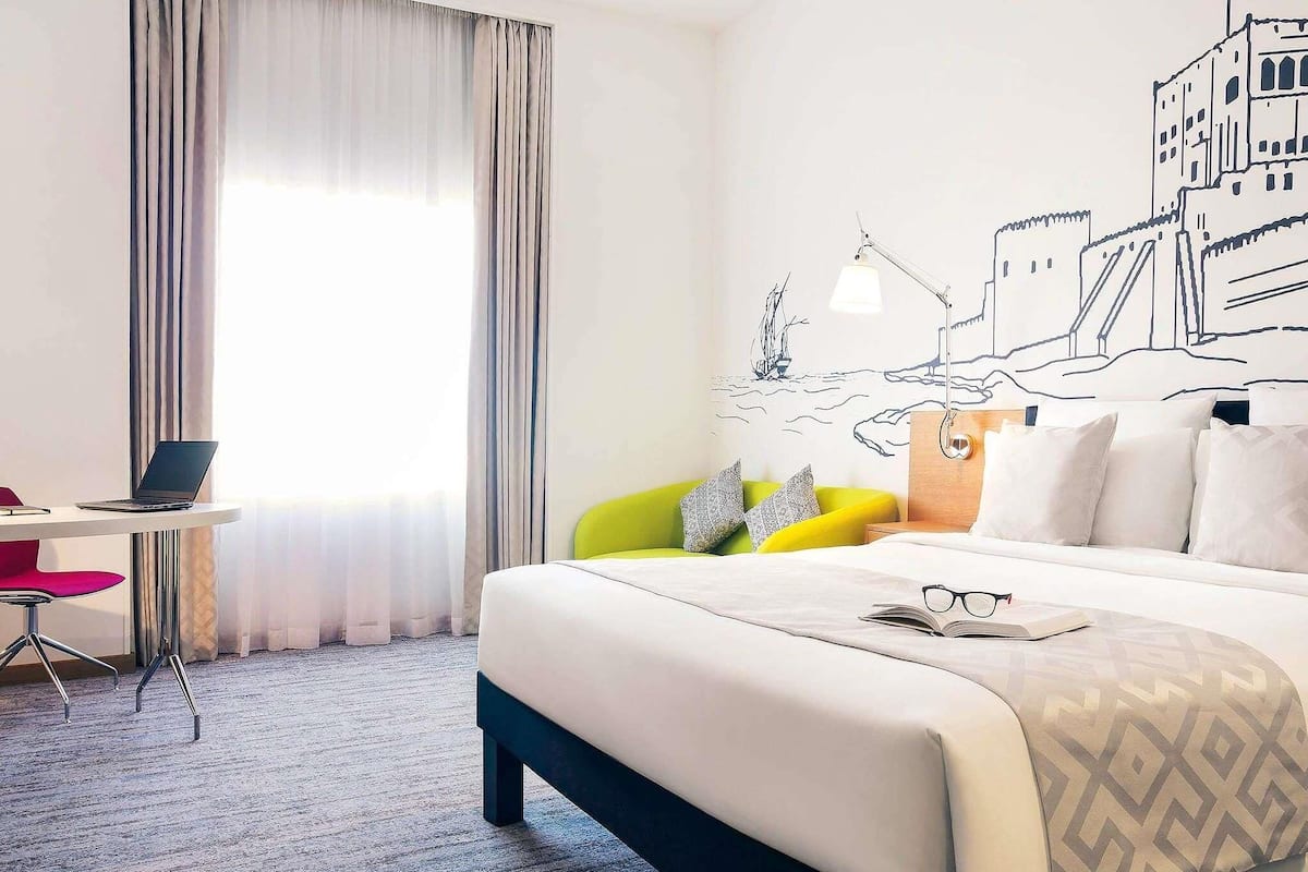 A hotel room with a bed, a desk with a laptop, a chair, and a wall mural.