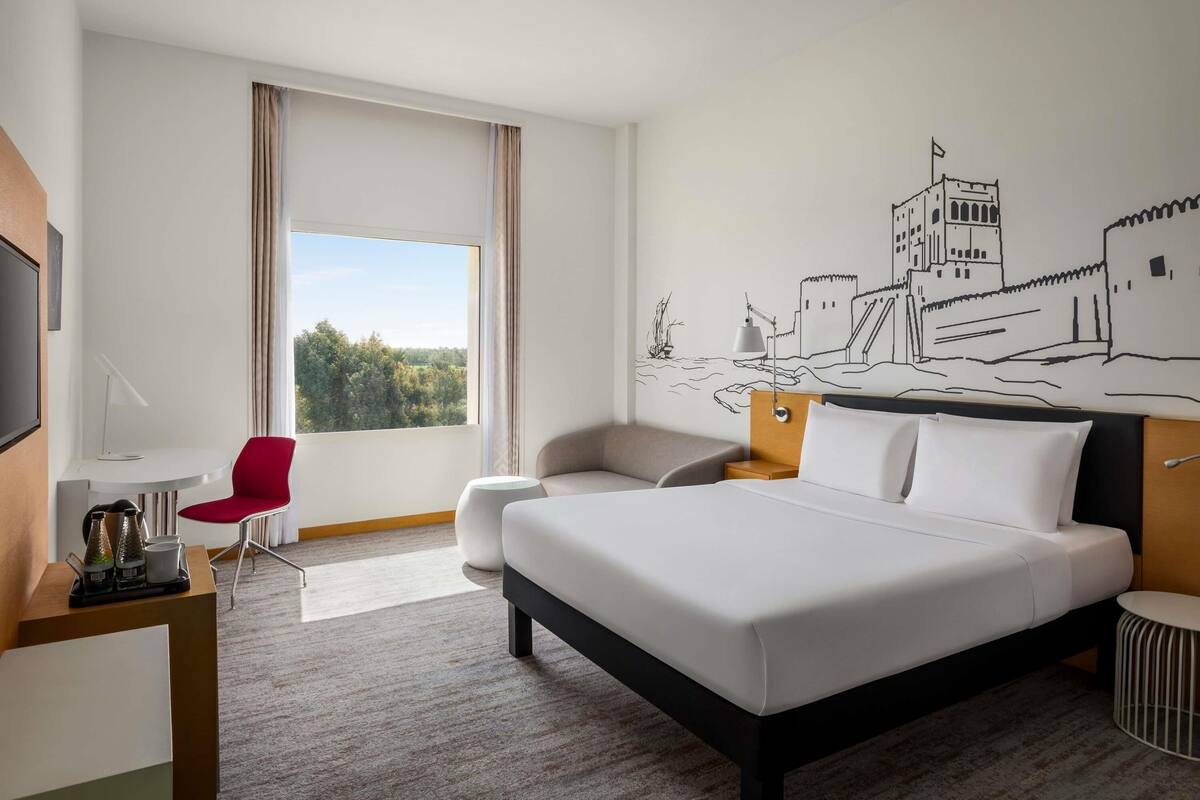 Una camera d'albergo con un letto, una scrivania con una sedia, una televisione e un'ampia finestra con tende.