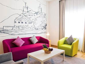 A living room with a vibrant pink sofa, a green armchair, and a white coffee table with a fruit basket. A large wall mural depicts a mountainous landscape.