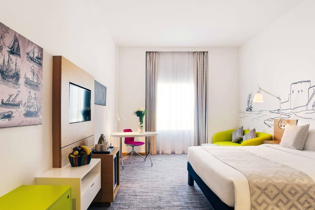 Una camera d'albergo con un letto, una scrivania con una sedia, un tavolino con un vaso e opere d'arte alle pareti.