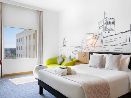 A hotel room with a bed, a bright green chair, a wall mural, and a large window.. Mercure Sohar