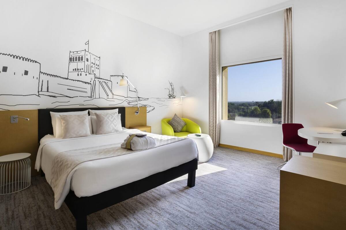 Una camera d'albergo con un letto, una scrivania, una sedia, una lampada e un'ampia finestra vista.