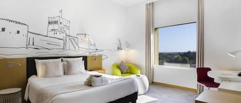 Habitación de hotel con cama, escritorio, silla, lámpara y un ventanal con vistas.
