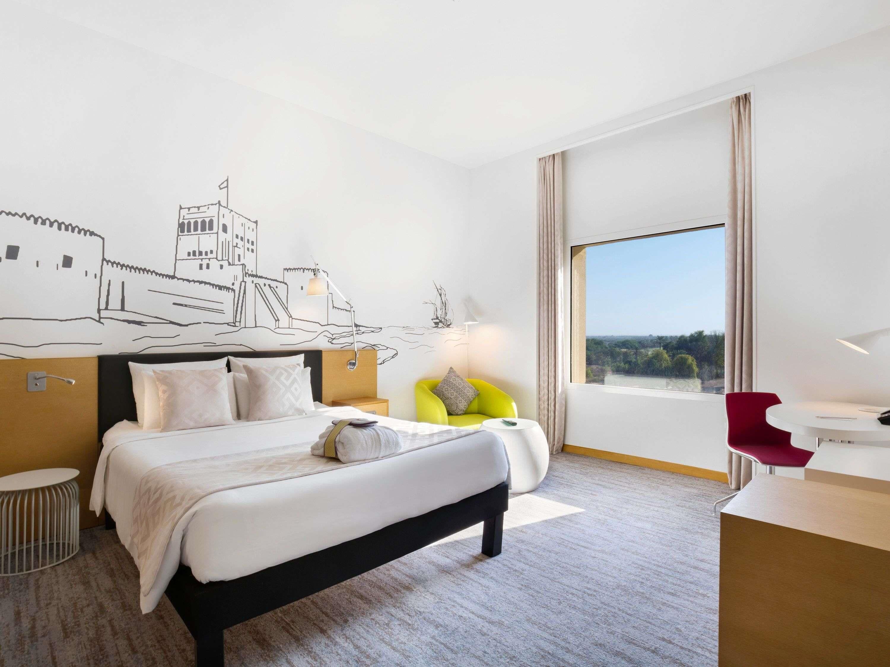 A hotel room with a bed, a desk, a chair, a lamp, a window with a view, and a wall mural.